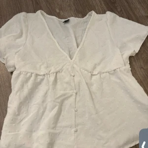 Vit blus med v-ringning och knappar - Söt vit blus med korta ärmar, v-ringad hals och dekorativa knappar framtill. Blusen har ett diskret prickigt mönster i tyget och lätt rynkade detaljer under bysten för en chill och feminin look. Perfekt att matcha med jeans eller kjol.