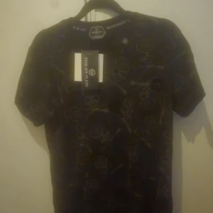 Svart t-shirt med döskalle från Philipp Plein - Svart t-shirt från Philipp Plein med Platinum Cut. T-shirten har korta ärmar och är täckt av gråa döskalle- och benmönster med PP-logga. Materialet är mjuk bomull och passformen är normal. Perfekt för dig som gillar edgy streetstyle.