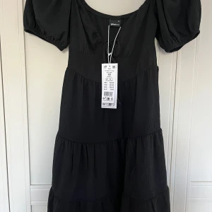 Svart puffärm klänning Gina Tricot XS - Säljer en svart kort klänning från Gina Tricot i storlek XS. Klänningen har puffärmar, knytning framtill och en volangad kjol. Materialet är lätt och har en diskret struktur som ger en cool vibe. Perfekt för dig som gillar trendiga och bekväma outfits.