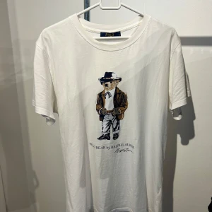 Vit Polo Bear t-shirt från Ralph Lauren - Vit t-shirt från Ralph Lauren med Polo Bear-tryck framtill. Björnen har cowboystil med hatt, jacka och boots. Rund halsringning och korta ärmar. Mjuk bomullskvalitet och klassisk passform. Perfekt för dig som gillar ikoniska prints och streetwear.