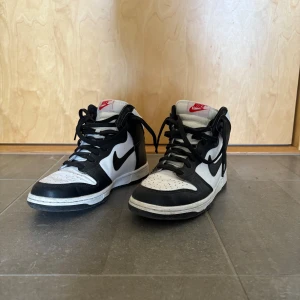 Nike Dunk High svart/vit sneakers - Säljer ett par Nike Dunk High sneakers i svart och vitt med klassisk röd Nike-logga på plösen. Skorna har snörning, rund tå och platt sula. Ovandelen är i skinn och de har en stilren, kontrasterande färgblock-design som ger en riktigt clean vibe.