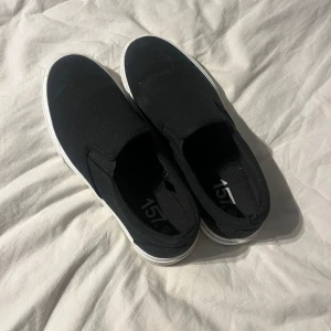 Svarta slip-on sneakers från lager 157 - Svarta slip-on sneakers från lager 157 med vit platt sula och rund tå. Skorna är tillverkade i bomullstyg och har en enkel, stilren design utan snörning. Perfekta för dig som gillar minimalistisk och clean streetstil.