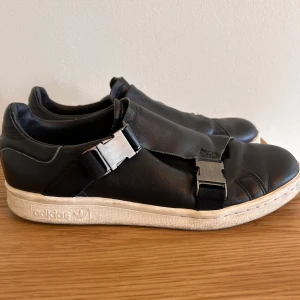 Adidas Stan Smith svarta sneakers - Svarta Adidas Stan Smith sneakers i skinn med coola metallspännen istället för klassisk snörning. Vit platt sula med Adidas-logga på sidan och Stan Smith-embossing på hälen. Stilren och unik design som sticker ut från mängden.