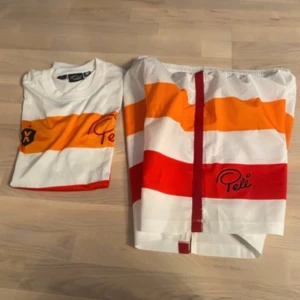 Randig Pelé fotbollströja L - Snygg fotbollströja från Pelé i storlek L med breda horisontella ränder i vitt, orange och rött. Tröjan har korta ärmar, rund hals och tryck med Pelé-logga samt emblem på bröstet. Materialet är lätt och andas, perfekt för match eller träning.