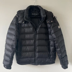 Svart pufferjacka från BVALDI - Säljer en svart pufferjacka från BVALDI med huva och coolt metallmärke på ärmen.                         (luva går att ta av)