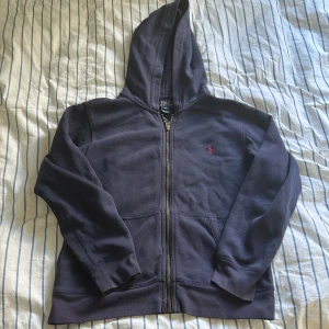 Mörkblå hoodie från Polo Ralph Lauren - Snygg mörkblå hoodie från Polo Ralph Lauren med klassisk röd broderad logga på bröstet. Tröjan har huva, dragkedja framtill och två praktiska fickor. Tillverkad i mjuk bomull, perfekt för chill dagar och sköna promenader. Den är i barnstorlek men passar mig som bär XS