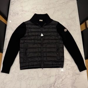 Svart dunväst från Moncler - Svart dunväst från Moncler med quiltad front och stickade ärmar. Klassisk ståkrage, dragkedja framtill och två fickor med dragkedja. Ikonisk Moncler-logga på vänster ärm. Perfekt för lager-på-lager och snygg streetstyle.