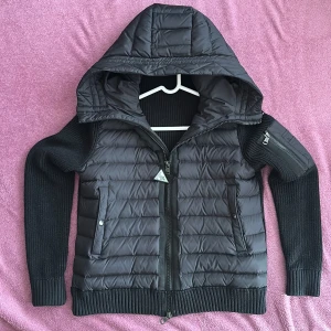 Svart Moncler cardigan  - Säljer en svart cardigan från Moncler med stickade ärmar och quiltad kropp. Jackan har en stor huva, dragkedja framtill, fickor med tryckknappar och en snygg Moncler-logga på ärmen. Perfekt för dig som vill ha en stilren och varm jacka med exklusiv känsla.
