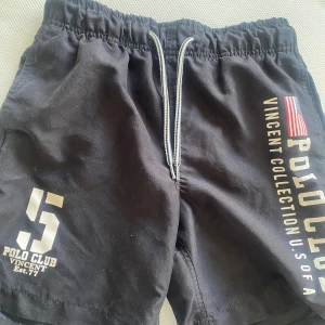 Svarta badshorts- Polo Club Vincent - Svarta shorts från Polo Club Vincent med tryck på båda benen, amerikansk flagga och siffran 5. De har snörning i midjan och en loose passform, perfekta för strand häng.. Materialet känns som polyester och de har sportig vibe.