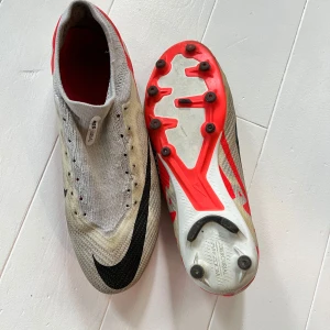 Nike Mercurial Air Zoom fotbollsskor - Nike Mercurial Air Zoom fotbollsskor i vitt med svarta och röda detaljer. Skorna har en strumpliknande ovandel i syntetmaterial och en platt sula med dobbar för konstgräs. Snyggt svart nike logga på tån och röda detaljer på hälen. Perfekt för dig som vill ha speed och kontroll på planen.