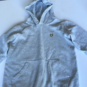 Grå hoodie från Lyle & Scott Junior - Snygg ljusgrå hoodie från Lyle & Scott Junior med klassisk gul fågel-logga på bröstet. Tröjan har huva, magficka och långa ärmar. Perfekt för chill dagar och enkel att matcha med jeans eller joggers. Pris går att diskuteras 