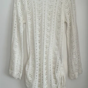 Vit stickad kortklänning med snörning - Supersöt vit stickad klänning med lång ärm och v-ringning. Klänningen har ett genomgående hålmönster och snörning i sidan som ger en snygg detalj. Har använt den som strandklänning, använd 1 gång. Storlek S