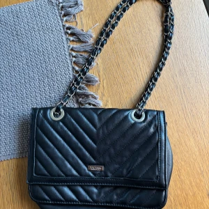 Svart axelväska från Don Donna - Snygg svart axelväska från Don Donna med kedjedetaljer på axelremmen. Väskan har ett lock med magnetknäppning, dragkedja på baksidan och flera fack inuti. Perfekt storlek för det viktigaste och ger en cool vibe till din outfit.