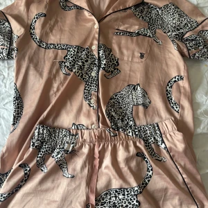 Leopardmönstrad pyjamasar från Victoria's Secret - Snygg pyjamas från Victoria's Secret i ljusrosa med svart och vit leopardmönster. Setet har skjortkrage, knappstängning, bröstficka med logga och elastisk midja med snörning. Materialet är mjukt och lent polyester, perfekt för chillkvällar. Ny pris 800kr