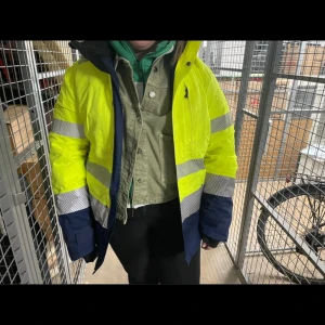 Gesto gul och marinblå pufferjacka - Säljer en varsel pufferjacka från Gesto Workwear i storlek L. Jackan är knallgul med marinblå detaljer och har breda reflexband runt ärmar och midja. Utrustad med huva och dragkedja, perfekt för dig som vill sticka ut och synas. Materialet är slitstark polyester.