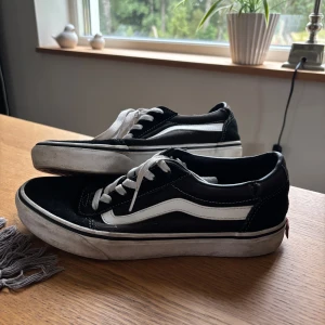Svarta Vans Old Skool sneakers strl 40 - Klassiska Vans Old Skool sneakers i svart och vitt med den ikoniska sidoremsan. Skorna har snörning, platt sula och rund tå. Ovandelen är i mocka och canvas, med vita kontrastsömmar och detaljer. Perfekt för dig som gillar streetstyle och skate. Sista bilderna visar att det finns lite skav på sidan. 