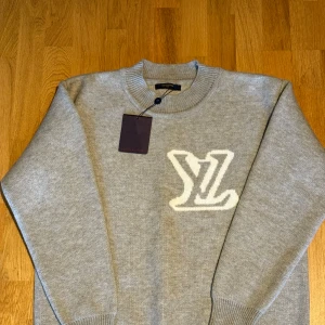 Grå stickad tröja Louis Vuitton - Supersnygg grå stickad tröja från Louis Vuitton som har prislappen kvar med stort vitt LV-logo på bröstet. Tröjan har rund hals och ribbade muddar vid ärmslut och nederkant. Perfekt för dig som gillar exklusiva märken och vill ha en clean look med lyxig touch. Säljs pga att den var för stor så aldrig använd