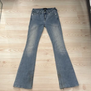 True Religion ljusblå bootcut jeans - Säljer ett par ljusblå jeans från True Religion med klassiska vita sömmar och snygga fickdetaljer med knappar bak. Modellen har bootcut och låg midja, tillverkade i jeansmaterial. Perfekt för dig som gillar retrovibbar och vill ha en cool siluett.