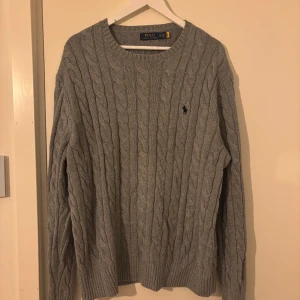 Grå kabelstickad tröja Polo Ralph Lauren - Säljer en grå kabelstickad tröja från Polo Ralph Lauren med klassisk rund hals och broderad logga på bröstet. Tröjan har långa ärmar och ribbade muddar vid ärmslut och nederkant. Perfekt för dig som gillar stilren och tidlös design. Helt ny aldrig använt, nytt pris 2250 kr