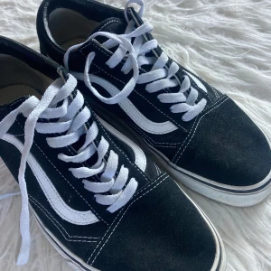 Vans svartvita sneakers - Svartvita Vans Old Skool sneakers med klassisk vit sidorand och vit snörning. Skorna har en platt sula och rund tå, tillverkade i mocka och canvas med kontrasterande vita sömmar. Perfekt för dig som gillar streetstyle och skate-vibe.