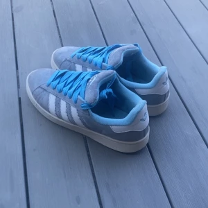 Adidas Campus sneakers grå/blå - Säljer ett par Adidas Campus sneakers i grå mocka med vita ränder och blå detaljer. Skorna har blå snören och blå insida, samt vit häl med Adidas-logga. Klassisk rund tå och platt sula, perfekt för dig som gillar retrostil och färgkombinationer.