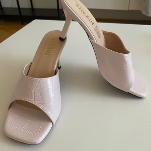 Ljusa rosa klackade sandaler från SHEIN - Säljer ett par ljusrosa sandaler från SHEIN med snyggt krokodilpräglat mönster. Skorna har öppen tå, fyrkantig framdel och smal hög klack. Materialet är syntet och de har en stilren, modern vibe som passar till många outfits.