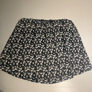 Svart blommig kortkjol  - Supersöt svart kortkjol från 157 med vitt blommigt mönster. Kjolen har en lätt utsvängd form och resår i midjan för skön passform. 