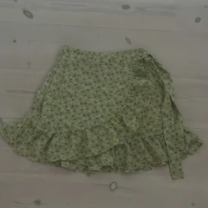 Blommig omlottkjol från FB sisters(New yorker)XS - Supersöt kort omlottkjol från New yorker i storlek XS. Kjolen är ljusgrön med blommigt mönster i lila, vitt och gult. Volangkant längs nederdelen och knytband i sidan ger en extra feminin touch. Perfekt för dig som gillar färggranna kjolar.