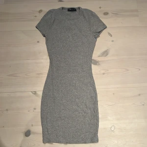 Grå långklänning FB Sister XS - Supersnygg grå ribbad lång klänning från FB Sister i storlek XS. Klänningen har en tight passform, rund hals och korta ärmar. Perfekt för dig som gillar enkel och stilren look med en kroppsnära siluett. Materialet är mjukt och stretchigt, vilket gör den bekväm att ha på.