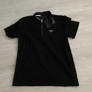 Prada  - Svart pikétshirt med klassisk krage och halv dragkedja framtill. Tröjan har ett diskret Prada-emblem på bröstet och är tillverkad i mjuk bomull. Perfekt för dig som gillar stilrena och exklusiva plagg.