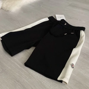 Moncler shorts  - Snygga svarta shorts med breda vita paneler längs sidorna och broderad logga på vänster ben. Modellen har elastisk midja med snörning för skön passform. Perfekta för chill eller sportiga dagar, stilrena och trendiga.