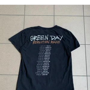 Green Day Revolution Radio t-shirt - Svart t-shirt från Green Day med Revolution Radio-turnétryck på ryggen och en brinnande boombox på framsidan. Rund hals och korta ärmar, tryck i vitt och orange. Perfekt för dig som älskar bandmerch och vill sticka ut med en cool grafisk design.