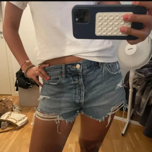 Blå jeansshorts från Zara med slitningar - Snygga blå jeansshorts från Zara med råa kanter och slitna detaljer. Klassisk femficksmodell med knapp och dragkedja. Perfekta för sommaren och ger en avslappnad vibe. Passformen är normal och shortsen är i ett mjukt denimtyg.