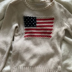 Beige stickad tröja North Sails M - Snygg beige stickad tröja från North Sails i storlek M. Framsidan har en stor broderad amerikansk flagga i blått, vitt och rött med grova stygn runt kanten. Tröjan är gjord av 50% ull och 50% nylon, har rund hals och långa ärmar. Perfekt för dig som gillar klassisk stil med en twist.