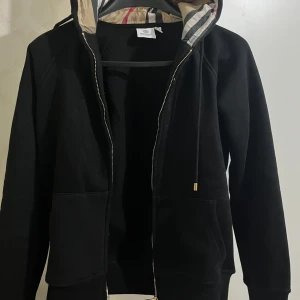 Svart hoodiejacka från Burberry - Svart hoodiejacka från Burberry med klassisk rutig insida på huvan, guldfärgade detaljer på dragkedja och snörning. Jackan har två stora fickor framtill och en snygg, stilren look. Perfekt för dig som gillar exklusiva streetwear-vibbar.