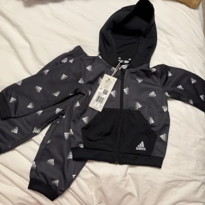 Baby Adidas tracksuit med huva - Snygg svart tracksuit från Adidas med huva och hel dragkedja. Setet har vita Adidas-loggor över hela materialet, ribbade muddar och elastisk midja på byxorna. Perfekt för dig som gillar sportig stil och vill sticka ut med klassisk design. Nypris: 650kr storlek: 6-9 månader.