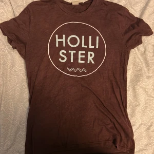 T-shirt Hollister - Snygg brun t-shirt från Hollister med stort vitt tryck på bröstet. Rund hals och korta ärmar, perfekt för chill dagar. Mjuk och skön bomull som känns lätt mot huden. Passformen är normal och t-shirten har en enkel, stilren look.