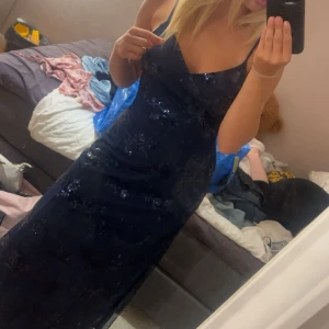 Marinblå glittrig balklänning  - Säljer en marinblå långklänning med tunna axelband och v-ringad front. Klänningen har glittriga paljetter över hela tyget och en öppen rygg som ger en snygg och festlig look. Materialet känns lätt och elegant, perfekt för speciella tillfällen.