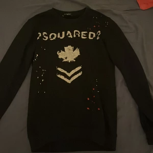 Svart Dsquared2 sweatshirt med print - Svart sweatshirt från Dsquared2 i storlek M men passar folk med S med coolt vitt och rött print framtill. Tröjan har lång ärm och klassisk rund hals. Materialet är mjukt och skönt, perfekt för dig som gillar streetwear med lite edge. Snygga detaljer med färgstänk och grafiskt tryck.