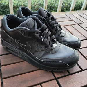 Nike Air Max 90 sneakers  - Säljer ett par svarta Nike Air Max 90 sneakers med klassisk design och synlig Air-dämpning i sulan. Skorna har snörning, rund tå och är tillverkade i skinn och syntetmaterial. Perfekta för dig som gillar streetstyle och vill ha en ikonisk sneaker i garderoben.
