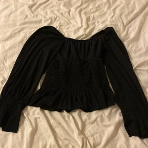 Svart offshoulder topp med volang - Trendig svart offshoulder topp med långa puffiga ärmar och smockad midja. Toppen har volangkant nedtill och är perfekt för dig som gillar en feminin och cool stil.