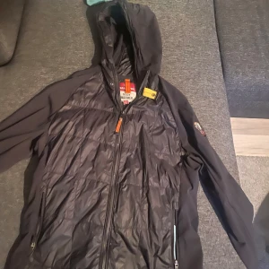 Svart jacka från Parajumpers - Nypris 4199kr mitt pris 999kr, pris kan diskuteras vid snabb affär. Svart jacka från Parajumpers med huva och dragkedja framtill. Perfekt för hösten och våren. Jackan har klassisk PJS-logga på ärmen och gul detalj vid halsen. Lättviktigt material med slät framsida och två fickor med dragkedja. Perfekt för dig som gillar stilren och sportig look. 