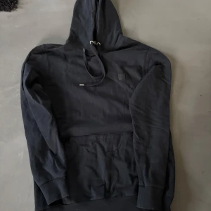 Svart hoodie från The Blue Uniform - Svart hoodie från The Blue Uniform i storlek M. Tröjan har huva med snörning och en stor magficka framtill. Klassisk och enkel design utan mönster, perfekt för chill dagar. Materialet känns mjukt och bekvämt, troligen bomull. Passar dig som gillar stilrena hoodies.