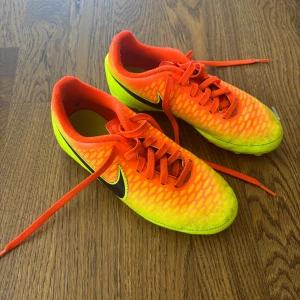Nike fotbollsskor orange/gul strl 33 - Säljer ett par grymma Nike fotbollsskor i en riktigt fet orange och gul färg med svart swoosh på sidan. Skorna har snörning och platt sula med dobbar för bästa grepp på planen. Ovandelen är i syntetmaterial med coolt mönster. Perfekt för dig som vill sticka ut på matchen! Ingen låda tyvärr
