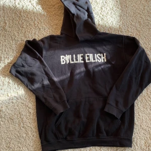 Svart Billie Eilish hoodie Fruit of the Loom - Svart hoodie med vit Billie Eilish-tryck på bröstet från Fruit of the Loom. Klassisk modell med huva och stor magficka. Mjuk bomullskänsla och långärmad, perfekt för dig som gillar streetwear och vill visa din musiksmak.