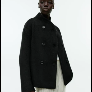 Svart ulljacja - Stilren svart ulljacja med dubbelknäppning och rak passform. Jackan har en hög krage och är tillverkad i ett ulligt material som ger en lyxig känsla. Perfekt för dig som gillar minimalistisk design och vill ha en jacka som sticker ut på ett snyggt sätt.