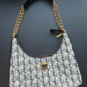 Quiltad axelväska från Guess - Snygg quiltad axelväska från Guess med guldfärgade kedjor och dragkedja. Väskan har ett rutigt mönster i svart, grått och beige med en  Guess-logga framtill. Materialet är en mix av ull. Väldigt elegant och fin!