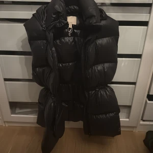 Svart lång dunväst med huva - Säljer en svart lång dunväst med stor, mysig huva och markerad midja. Västen är quiltad och har en glansig finish som ger en cool look. Perfekt att slänga över en hoodie för extra värme. Stängs med dragkedja framtill.