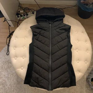 Svart dunväst med luva och dragkedja - Svart väst med luva. Dragkedja och även två fickor. Sidorna är ribbade för att forma passformen. Superbra skick, inga hål eller fläckar. Varm o skön, passar även Xs💗