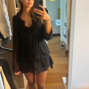 Svart kort volangklänning - Säljer en svart kort klänning med volangdetaljer vid halsringningen och nederkanten från Saint Tropez. Klänningen har långärmade transparenta ärmar och ett flowy, lätt material som ger en snygg och luftig look. Perfekt för dig som gillar romantiska detaljer och en chic stil.